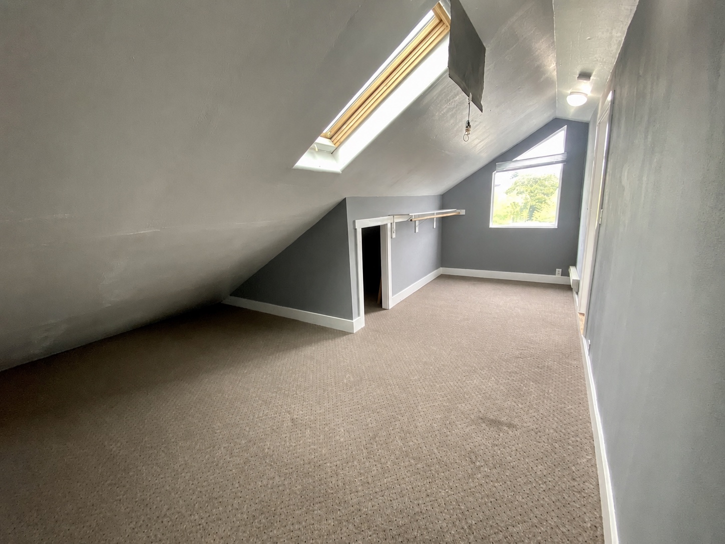 Attic bedroom before editing — cool grey walls reading slightly washed-out, skylight bright but flat, dormer window underexposed against the bright sky, beige carpet flat through the room.