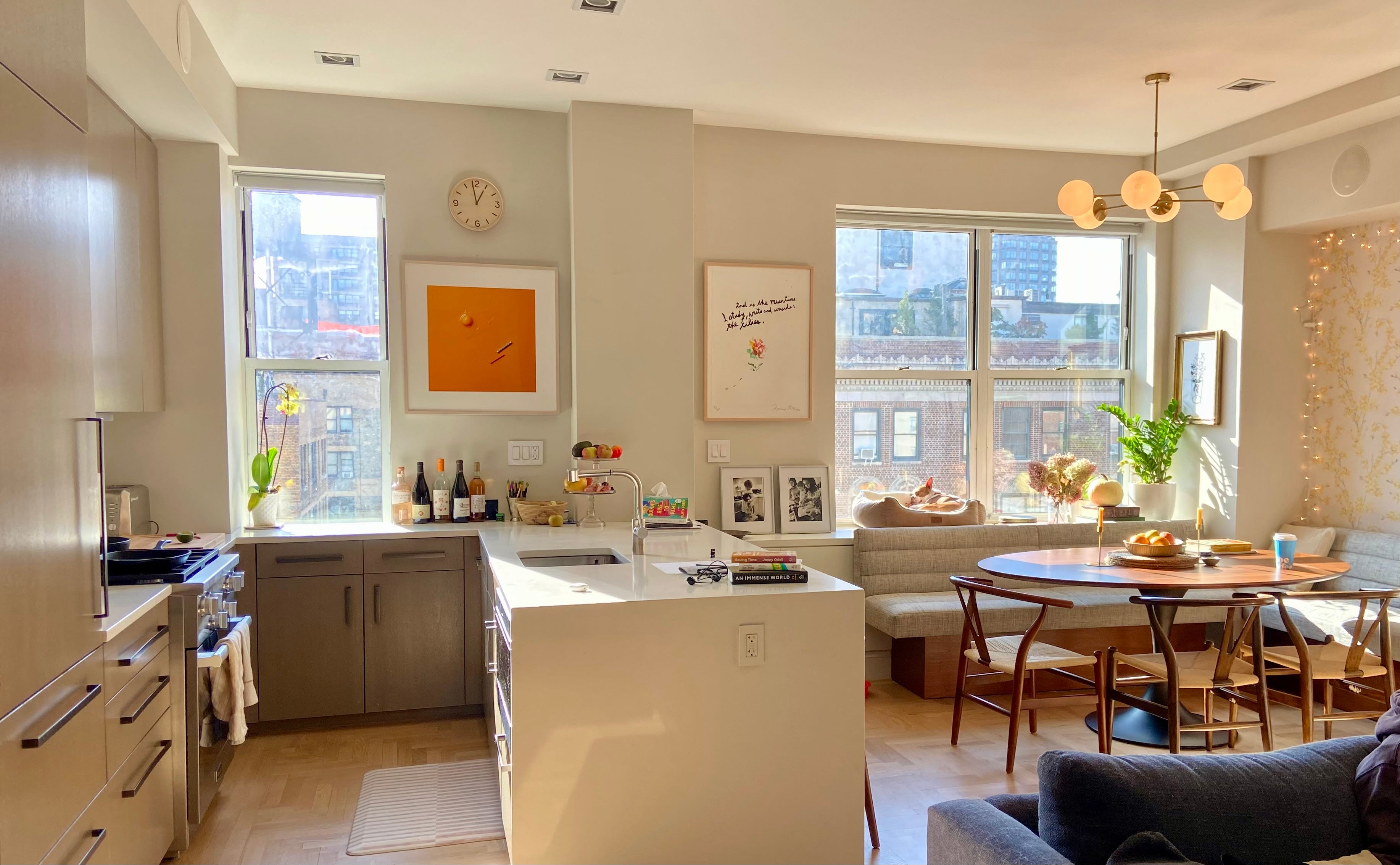Kitchen + dining before editing — warm-yellow cast across walls and ceiling, countertop clutter (wine bottles, books, frames, loose objects), pet bed and personal items on the bench.